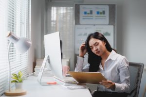 Young asian businesswoman looking stressed while working at her desk in modern office, holding a pen and reading notes on a clipboard