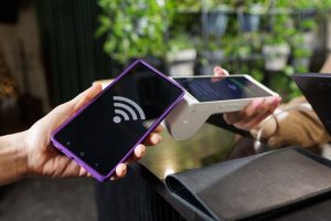 Customer uses smartphone NFC tap to pay on handheld POS reader at cafe counter, cashless checkout concept